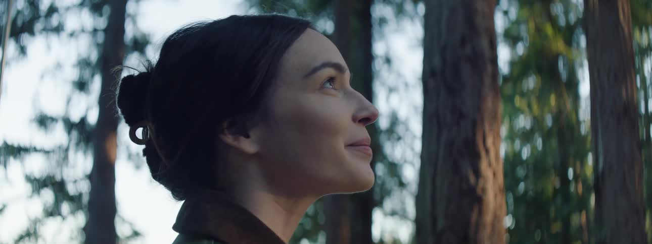 A woman smiles as she looks up at tall trees in the forest