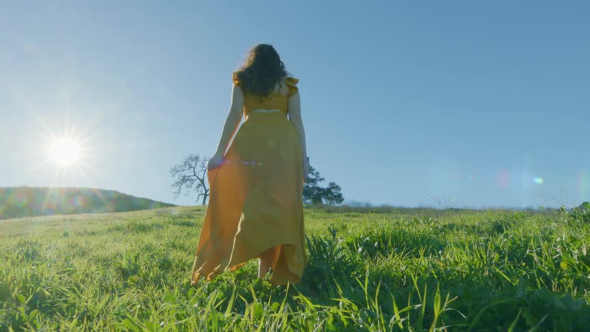 Back of a lady in yellow gown walking barefooted on grass at midday