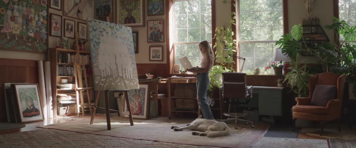 Woman standing using her laptop in a room with paintings and a white dog lying next to her