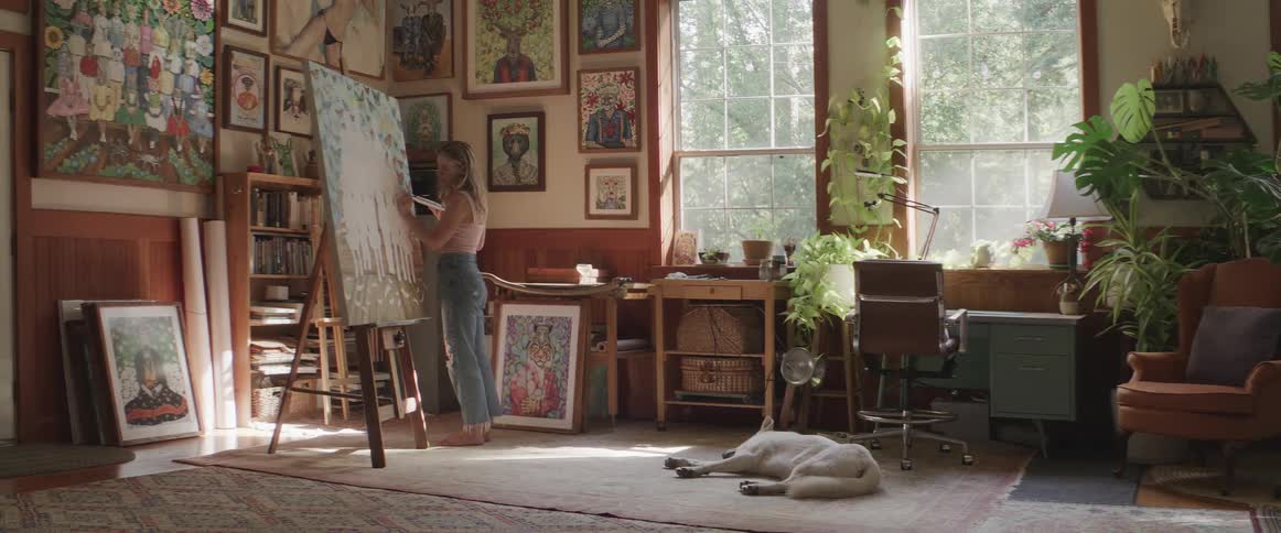 A woman painting in her living room with a dog lying on a rug on the floor
