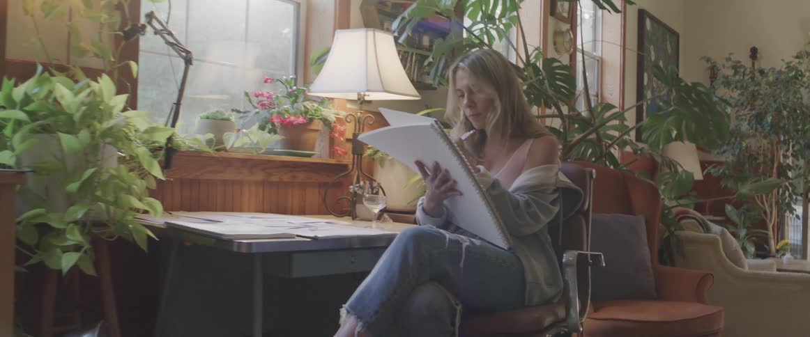 Blonde woman sits at desk indoors and holds large sketchbook