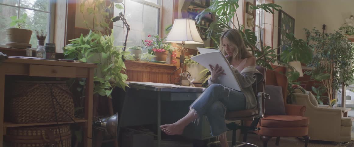 Woman sitting on a chair writing in a room with bushes and right in front of her a dog that is lying down sleeping