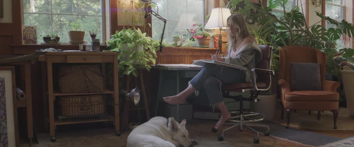 Woman sitting in a chair in the middle of a room drawing and her dog next to her sleeping