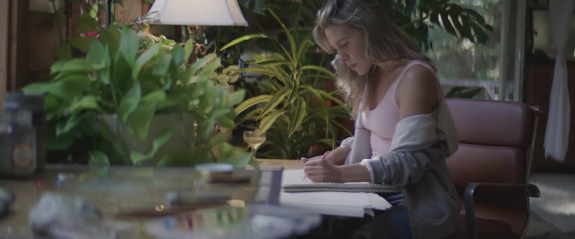 Woman sits at table in room decorated with multiple plants and sketches