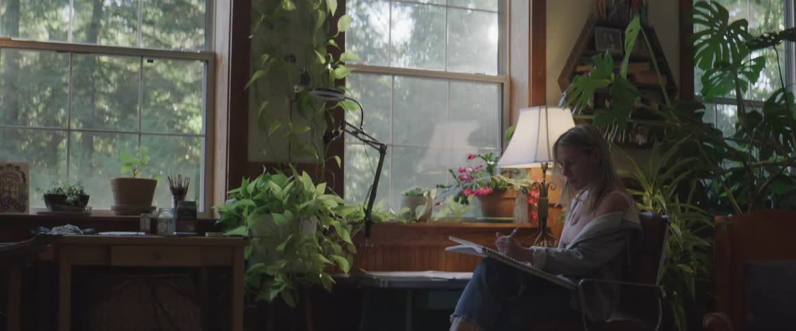 Woman sitting at a desk and writing in a large notebook inside a room with bushes
