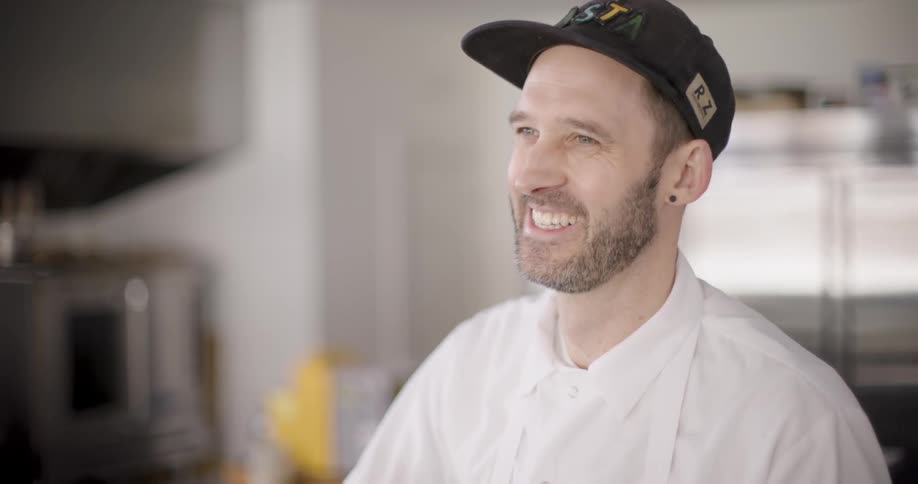 A man wearing a white shirt and black baseball cap laughs