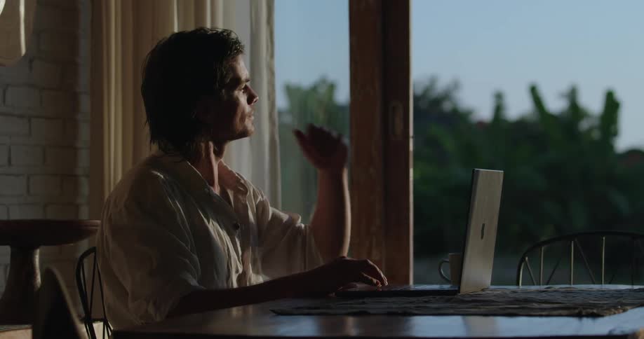 A man sitting at a table with a laptop looking out the window before he turns back to the laptop to write
