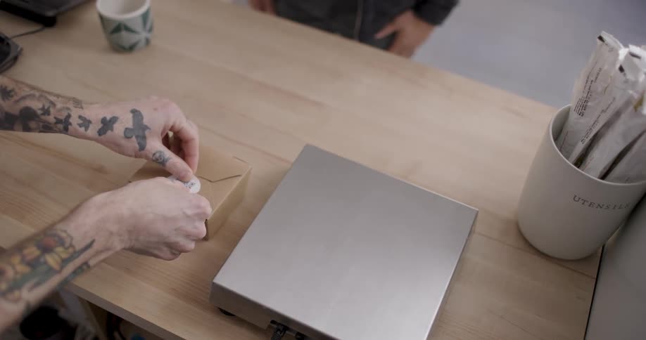 A person with tattooed arms places a sticker on a box over a wooden table