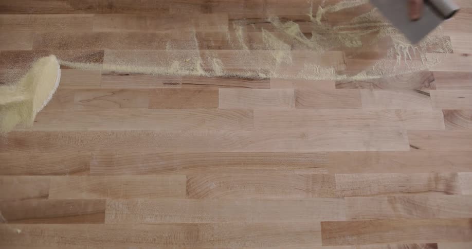 A persons hand with a spatula on a wooden countertop gathering flour