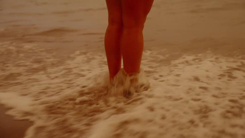 A woman stands on a sandy beach as a small waves washes over her feet