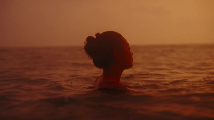 A young Asian woman is floating in the ocean with a dramatic orange sky in the background