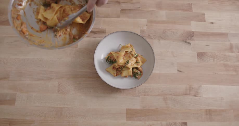 A person is serving pasta from a pan onto a white plate on a wooden surface