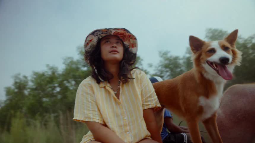 A young asian woman and her dog roll by on the back of a cart being pulled down a gravel road through the Philippines countryside