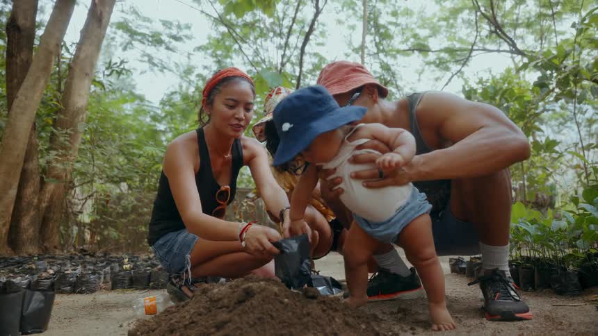 A couple in the woods holds a baby so it takes a seed out of a small plastic bag