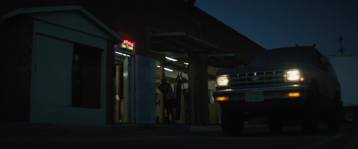 Three men practice boxing in a garage with its doors open and a pickup truck waiting outside