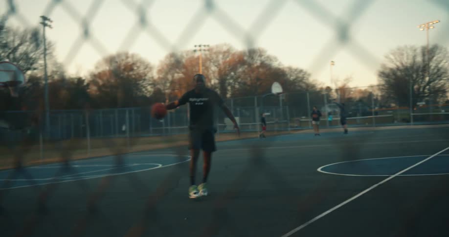 A man is hitting a basketball on the ground passing it between his legs and then throwing it towards a fence
