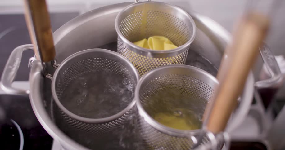A person cooks noodles in three miniature strainers inside a pot of boiling water