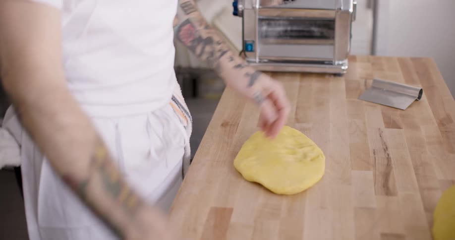 Hands of a tattooed man wearing an apron and kneading dough and adding flour