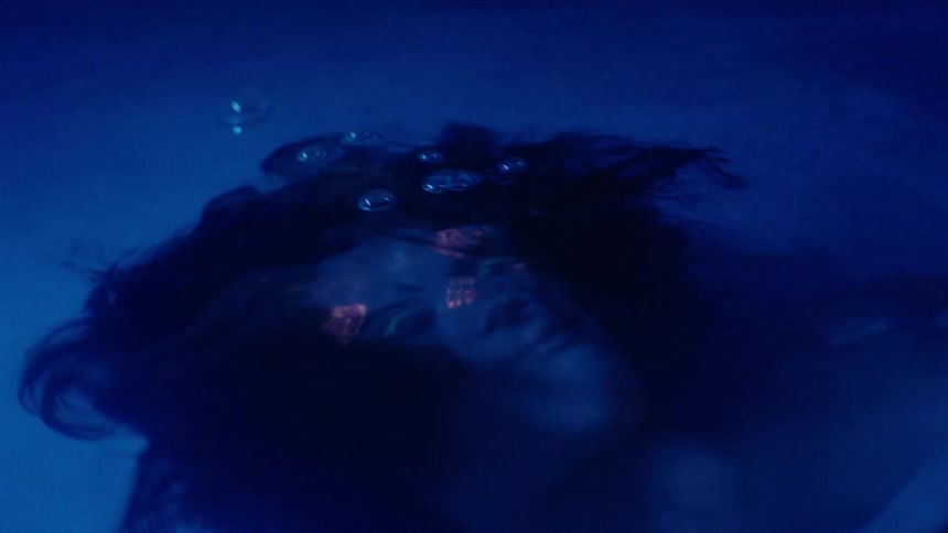 A young Asian woman with bright face paint submerges under water lit by blue lighting