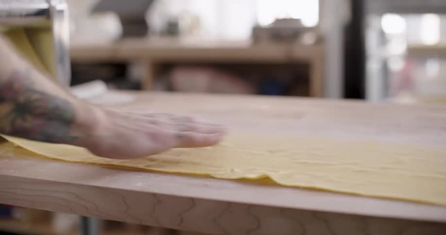 A person is using a pasta machine to roll out a sheet of fresh pasta on a wooden table in a welllit room