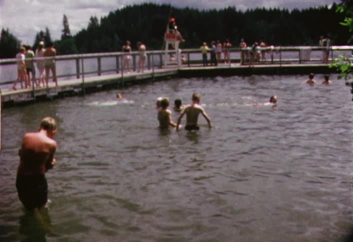 Several people splashing and swimming  in a lake as others walk across a low bridge