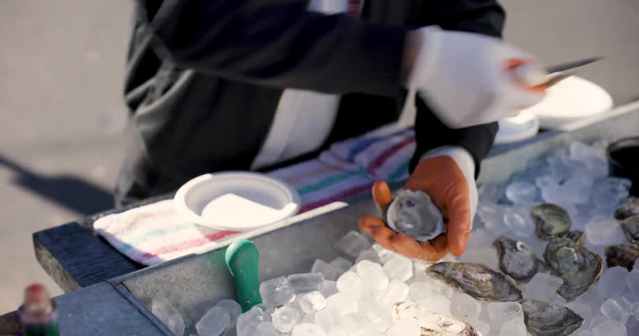 A person wearing gloves is using a knife to open oysters that are in a container with a lot of ice
