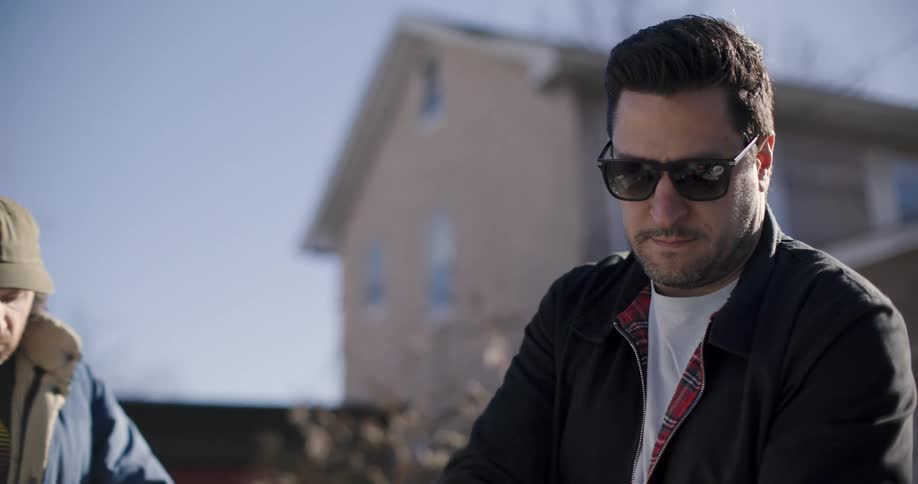 A man in a black jacket and sunglasses is preparing some food and behind him is a man who is eating