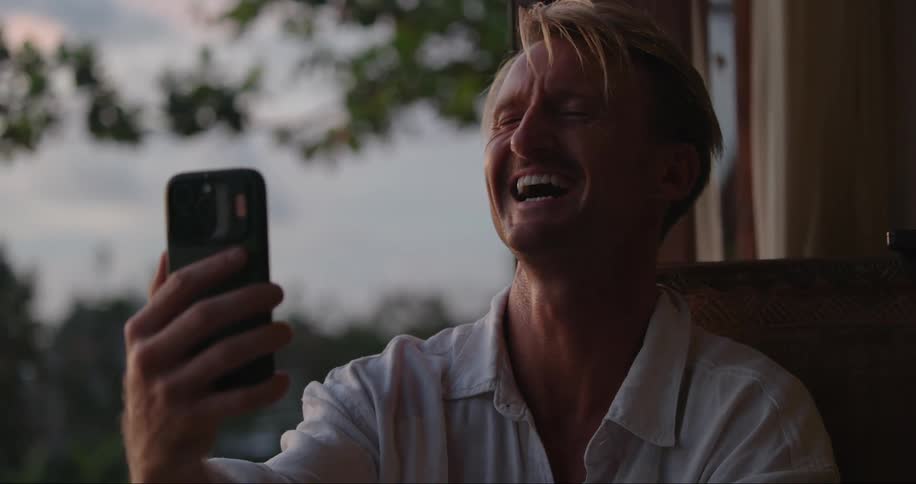 A man sits on his porch laughing as he holds his cell phone
