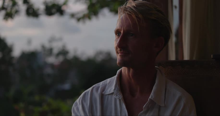 A man sitting on his porch hold his cell phone and blows a kiss as he talks then sits looking around