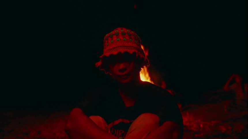 A young Asian woman wearing a patterned hat is sitting in front of a campfire