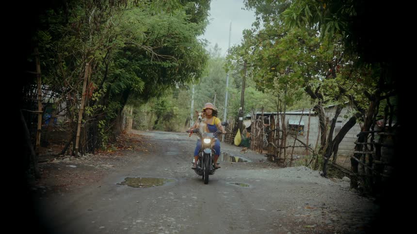 A young Asian woman and a dog ride by on a vespa motorcycle in a rural environment