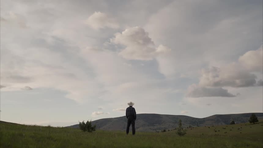 Man with cowboy hat on a post overlooking mountains on a sky day with clouds