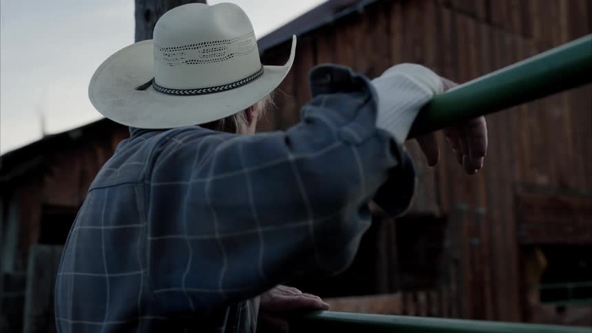 Elderly man wearing a cowboy hat leaning against a railing next to a wooden barn during the day