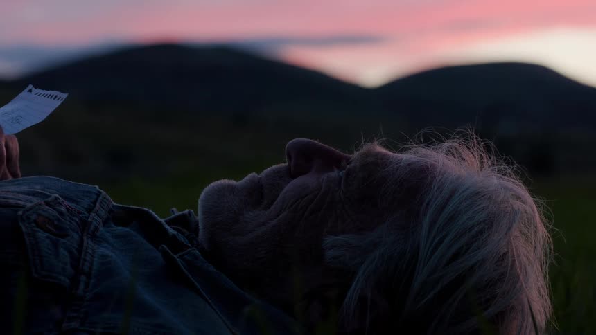 An old man with a piece of paper lies down outdoors