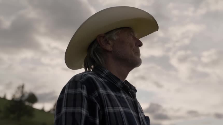An elderly man wearing a cowboy hat stands and thinks on a grassy hill