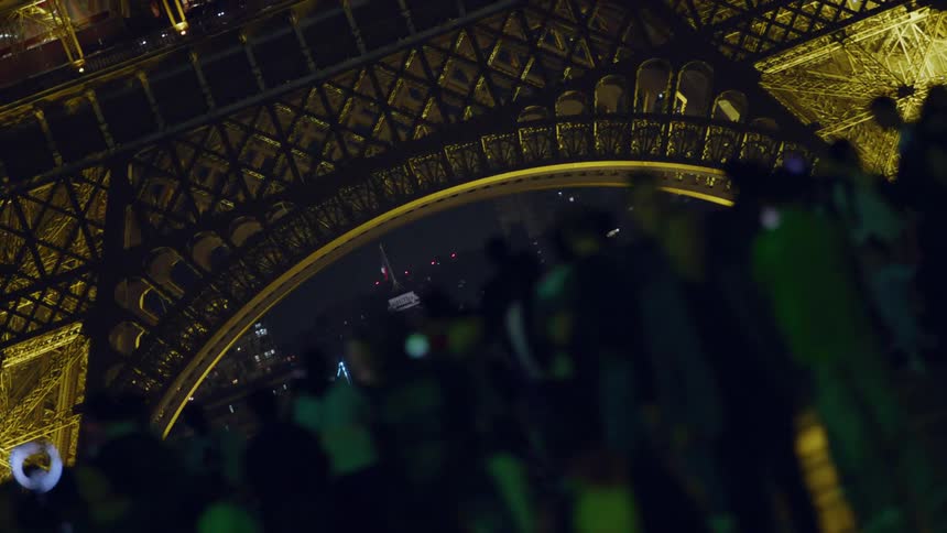Some people in the crowd at the base of the eiffel tower at night hold up smartphones