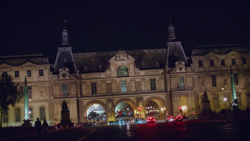 A large building with old architecture in front of a street with cars passing by and people too