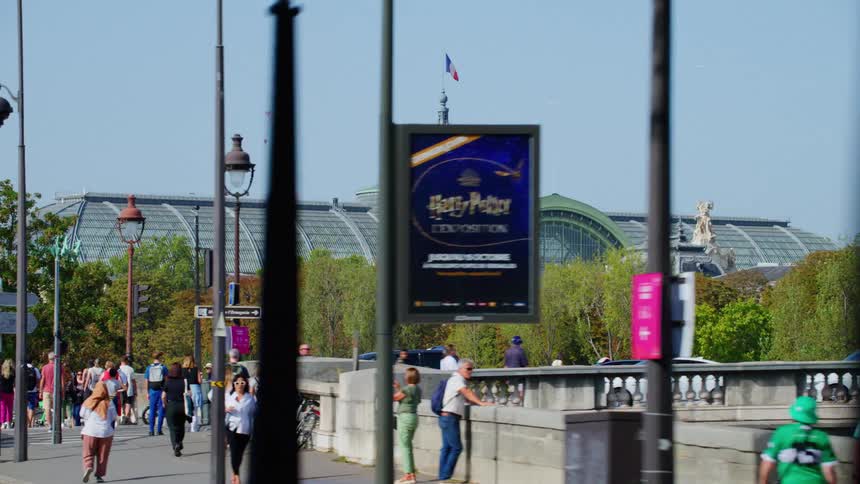 Pedestrians walk along wide sidewalk leading to long landmark building with arched roof