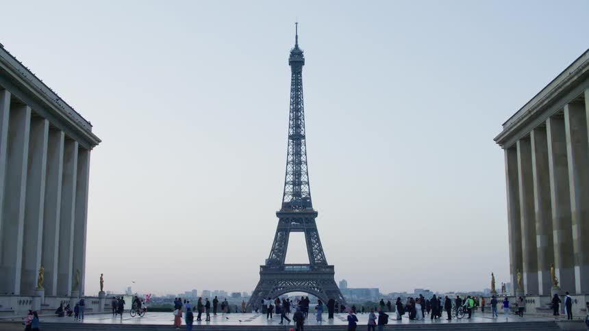 The eiffel tower stands tall between two large buildings with people walking around
