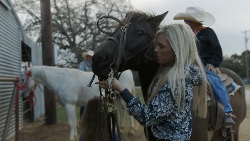 A boy is sitting on a horse as a woman tries to put its bridle on