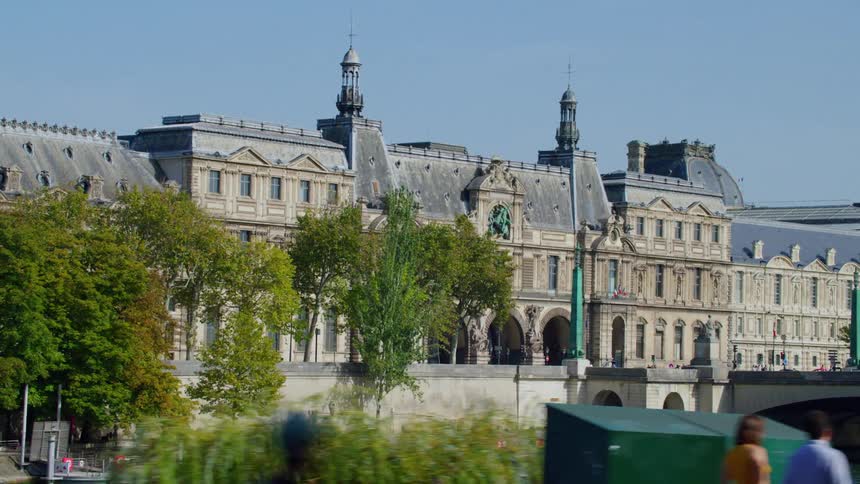 A large building with ancient architecture and huge doors facing a bridge that passes over a river and there are several people around
