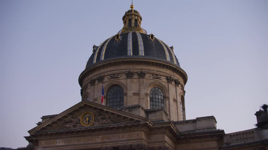 Outdoor clock sits under roof near base of the domed second story of a large extensive building