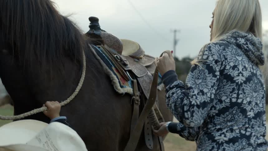 Woman removing a saddle from a horse as a child holds its reins