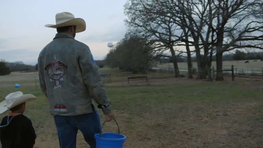 Man and boy  wearing cowboy hats walking through a pasture that has some horses in its and the man is carrying a bucket