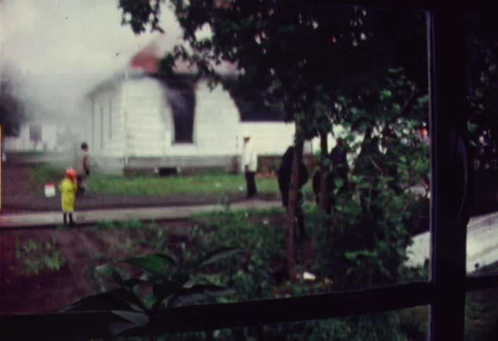 Smoke pours from the windows of a house as onlookers mill about