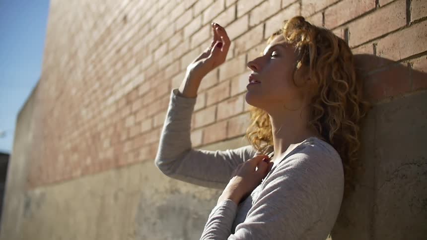 Woman leans against a brick building with the sun shining down on her
