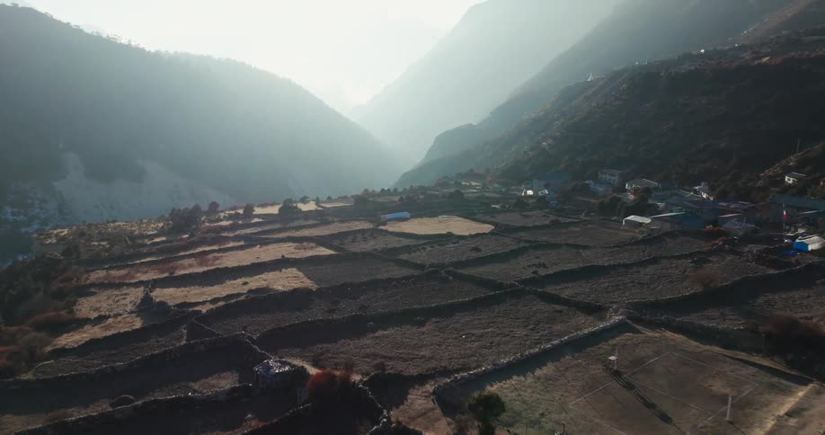 An aerial view of farmlands and a village illuminated by the bright sun shining over high mountains