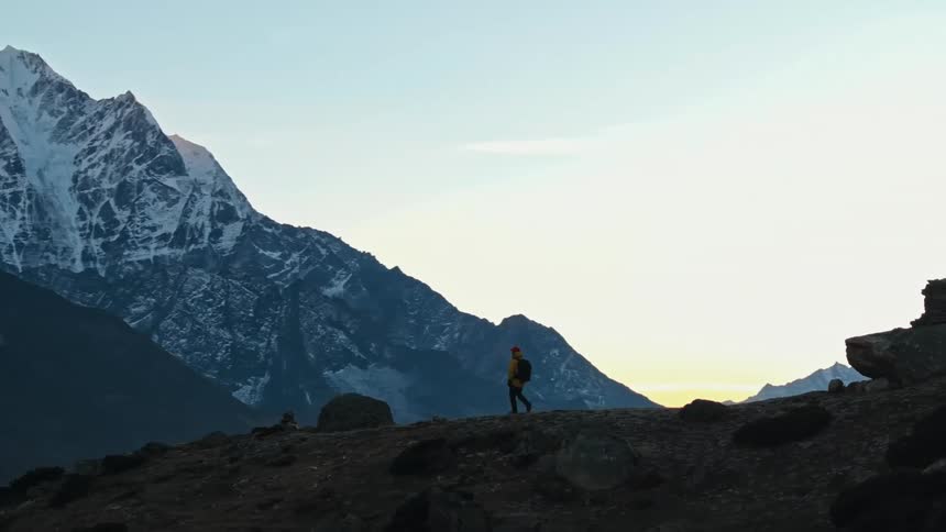 A person in a yellow jacket and a large backpack is walking in a region with many mountains and the tops are covered in snow