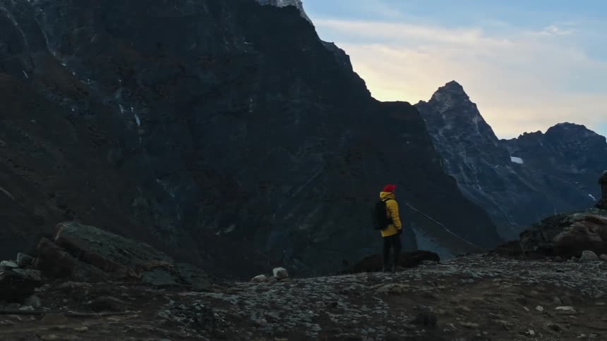 A hiker wearing a red hat a yellow coat and a black backpack lifts his hands up as he gazes at a distant high mountain range