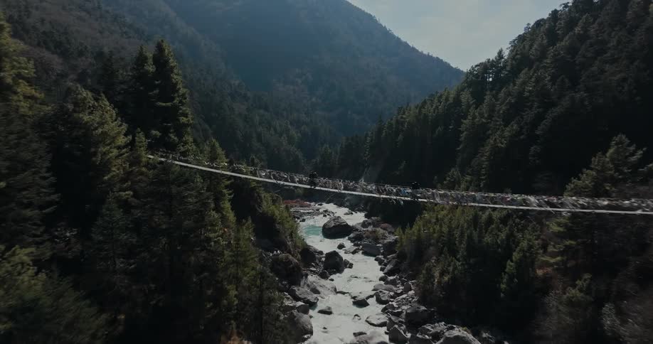 A river passes through a valley lined with forests as hikers cross a rope bridge
